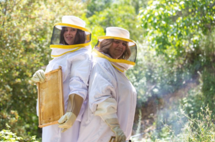 Une mère et sa fille debout, dos à dos, portant des tenues apicoles. La fille, à gauche, tient uncadre de ruche.