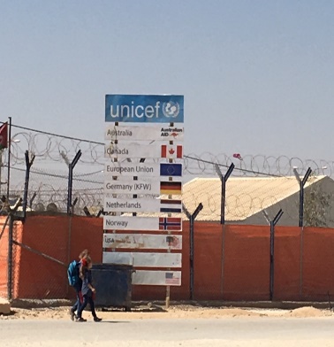 Image de deux personnes marchant sur le bord d'une route, devant une grande affiche de l'UNICEF portant les noms et les drapeaux de divers pays.