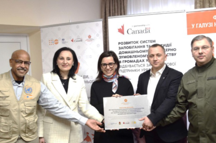 A group of 3 men and 2 women, including Canada’s former ambassador to Ukraine, Larisa Galadza, hold a certificate. There are signs placed directly behind them.
