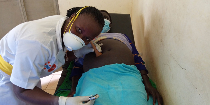 A midwife wearing a mask holds a Pinard horn to the abdomen of a pregnant woman, who is also wearing a mask.