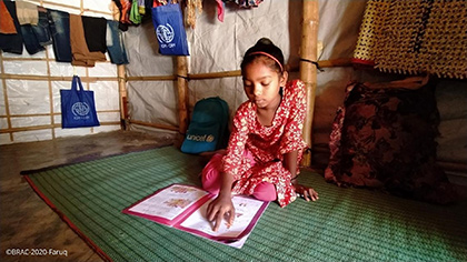 Une fille est assise par terre dans un abri et lit un manuel scolaire. Derrière elle se trouve un sac contenant l’inscription <q>« UNICEF »</q> et des vêtements accrochés aux tiges de bois qui forment les murs de l’abri.