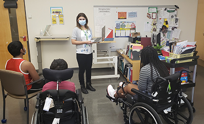 A teacher wearing a mask stands at the front of a classroom before three students. Two of them are sitting in wheelchairs and one is sitting on a chair.