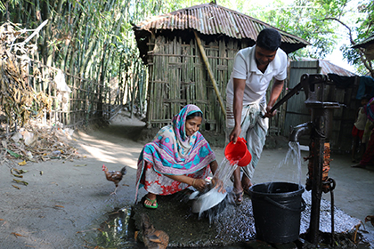Une femme et un homme tiennent un pot et un seau afin de récupérer l'eau qui s’écoule d'une pompe.