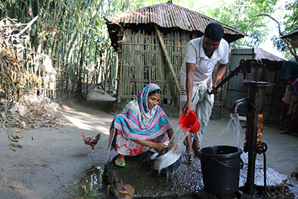 A woman and a man hold a pot and a pail under water pouring from a pump.