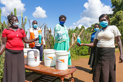 Four women and a man wearing masks show the liquid soap they made and put into bottles and large pails.