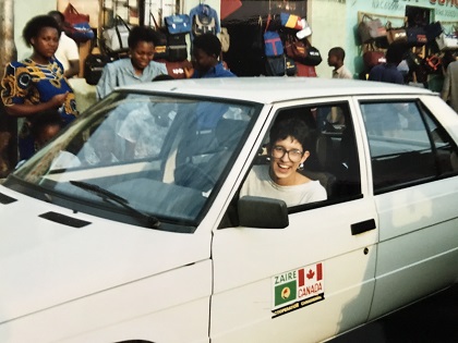 Isabelle Bérard souriante, au volant d’une voiture blanche sur laquelle sont apposés le drapeau du Canada et celui du Zaïre. En arrière-plan, des gens dans la rue..