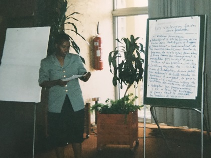 A woman from Rwanda, papers in hand, in front of a paper pad on which is written several sentences.