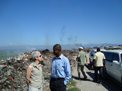 Isabelle Bérard in Haiti, speaking with a man. In the background are mounds of debris, smoke, and people walking in the street.