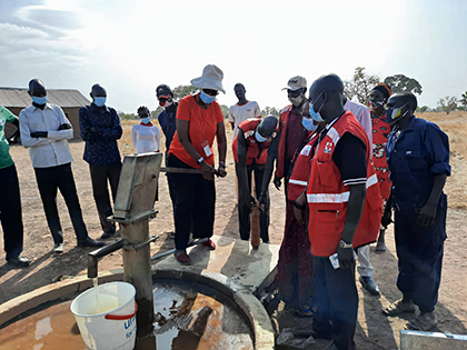 A dozen people are gathered around a water hand-pump