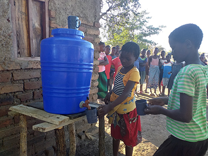 A child fills a cup with water from a dispenser. Several other children watch her do it.