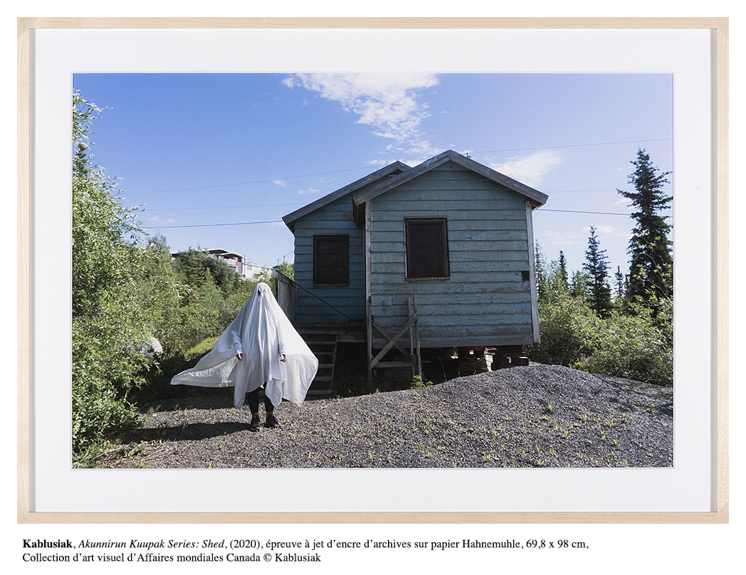 Une personne recouverte d’un drap blanc se tient devant une petite maison de bois de teinte bleutée.