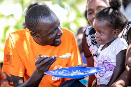 A man wearing an orange shirt smiles to a little girl with a plate and spoonful of food.