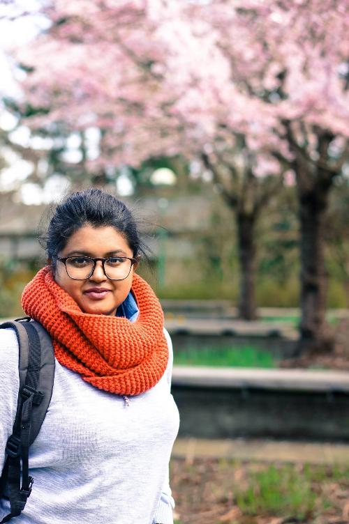 A woman wearing a grey shirt and an orange scarf looks into the camera; 2 cherry blossom trees can be seen in the background.