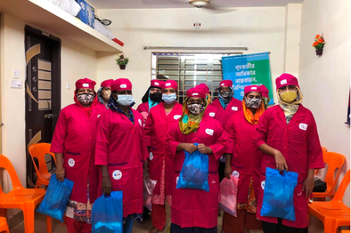 A group of women stand facing the camera. The women are wearing deep pink uniforms and holding plastic bags.