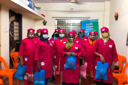 Un groupe de femmes se tient face à la caméra. Elles portent un uniforme rose foncé et tiennent un sac en plastique.