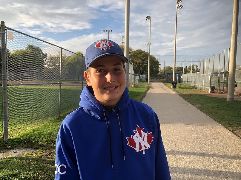 A boy wearing a blue hat and hoodie smiles in a baseball park.