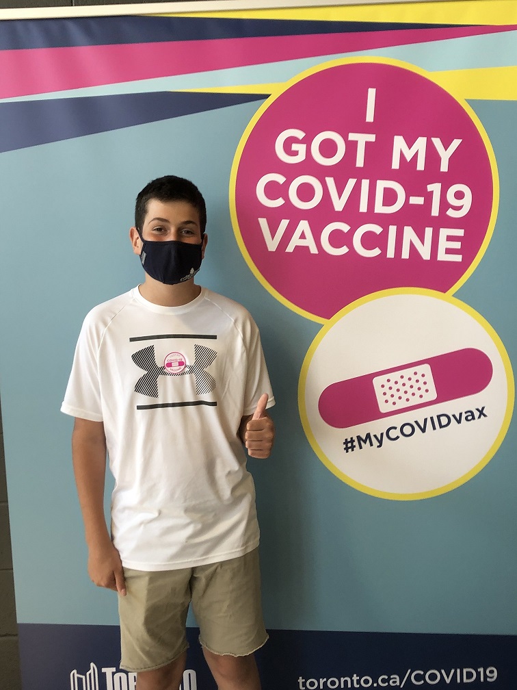 A boy wearing a mask gives a thumbs up in front of a poster that reads “I got my COVID-19 vaccine. #MyCOVIDvax.”