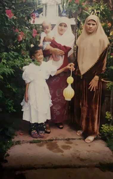 A young girl wearing a white dress holds the hand of another woman wearing a gold headscarf next to another woman wearing a red dress and white headscarf with a baby in her arms.