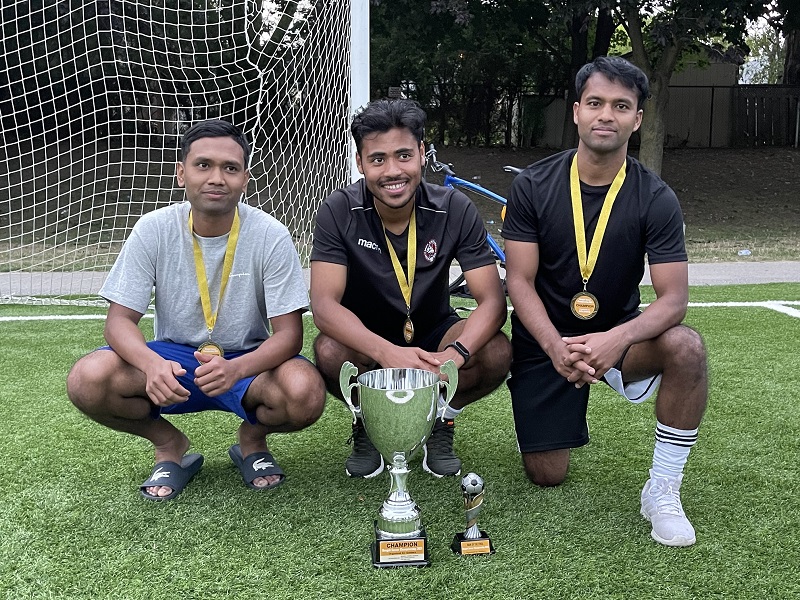 Three men smiling and wearing medals with yellow ribbon sit in front of a trophy in front a goal post.