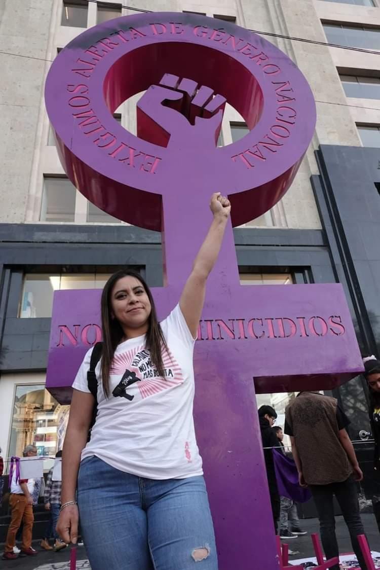 A Mexican woman stands with her fist raised in the air. Behind her is a symbol of Venus sculpture that has a raised fist in the centre of it.