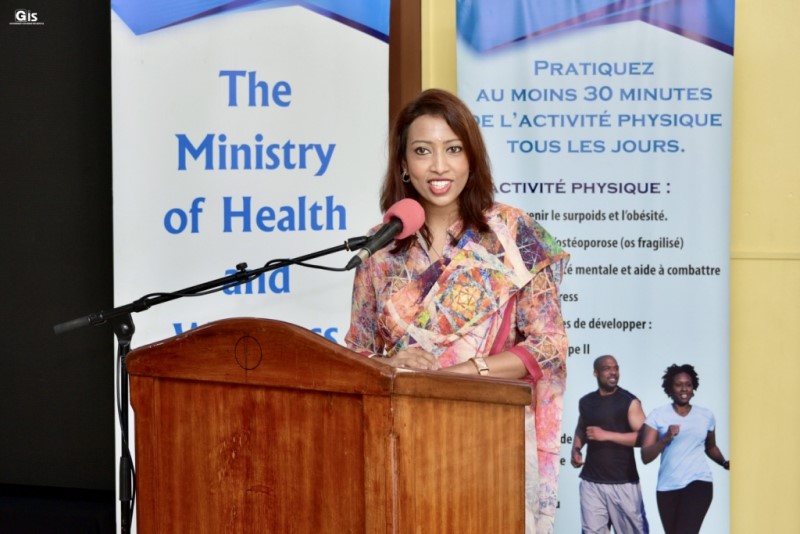 A Mauritian woman stands at a podium. Behind her are two banners for the Ministry of Health and Wellness.