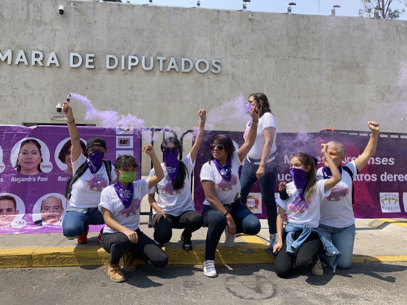 A group of Mexican girls, all wearing matching t-shirts and purple handkerchiefs over their faces or around their necks. Many have their fists raised.