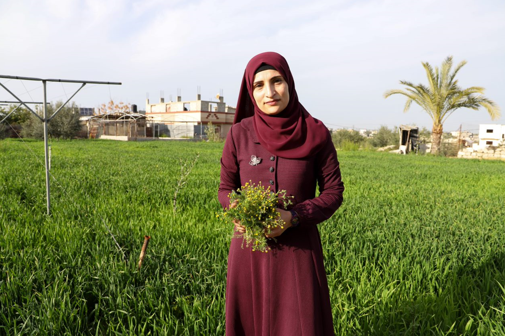 woman wearing a head scarf stands in a field and holds a bouquet of flowers.