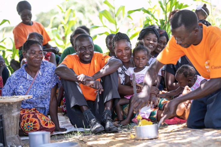 A group of smiling people watch as a man, kneeling on the ground, mixes food in a pot.