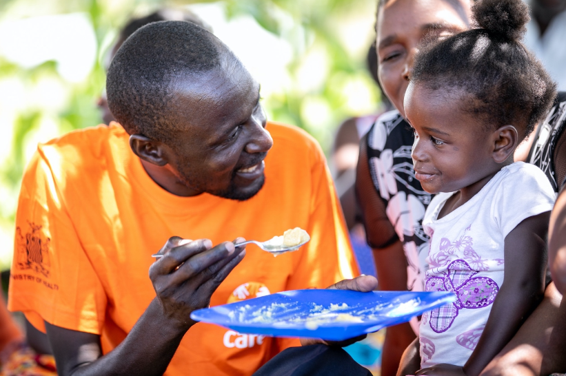 A man, holding a plate, smiles at a young child and offers them a spoonful of food.