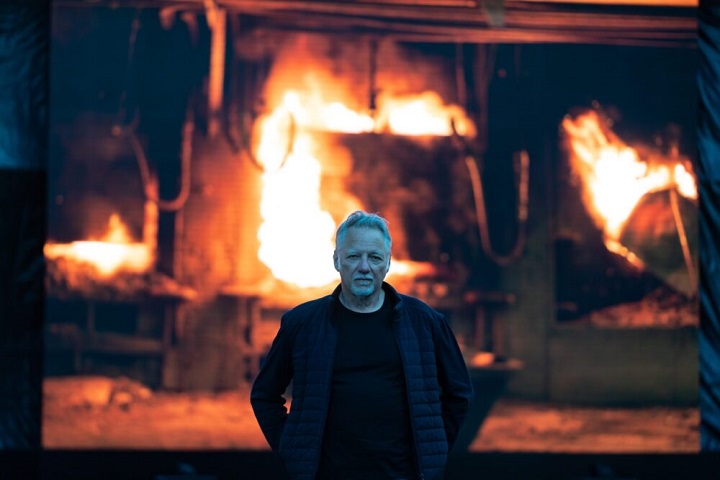 Edward Burtynsky debout devant une grande télévision montrant une image d'une pièce en flammes