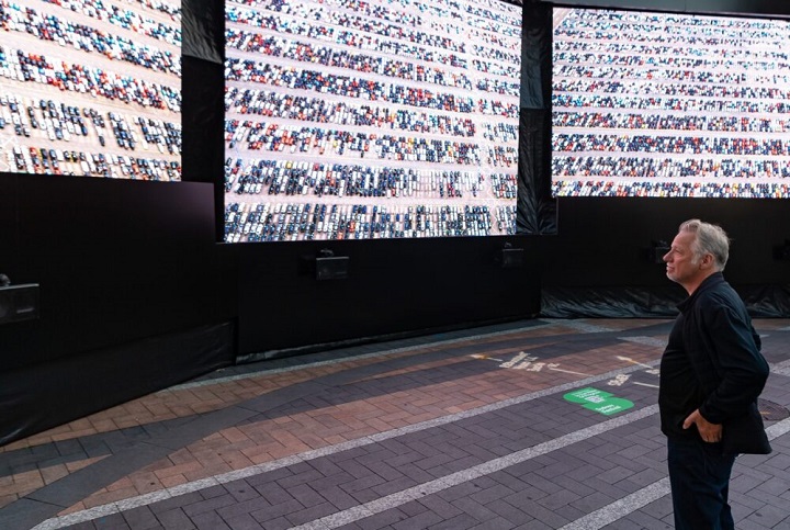 Edward Burtynsky debout devant trois grands téléviseurs montrant chacun des images de parkings remplis.