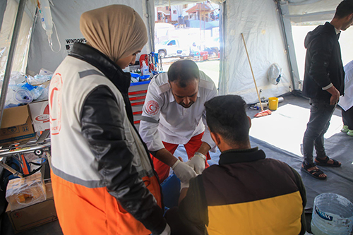 A woman in a Red Crescent Society vest watches while another Red Crescent Society paramedic performs a procedure on a man sitting in a chair