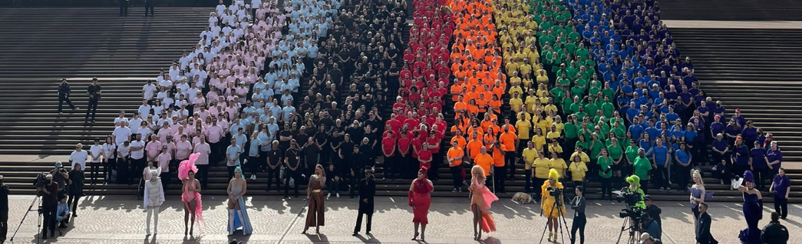 Canadian high commission staff celebrating Pride season in Australia. Diplomatic staff wearing solid colour shirts grouped by colour to symbolize a Pride flag in front of Sydney Opera House.