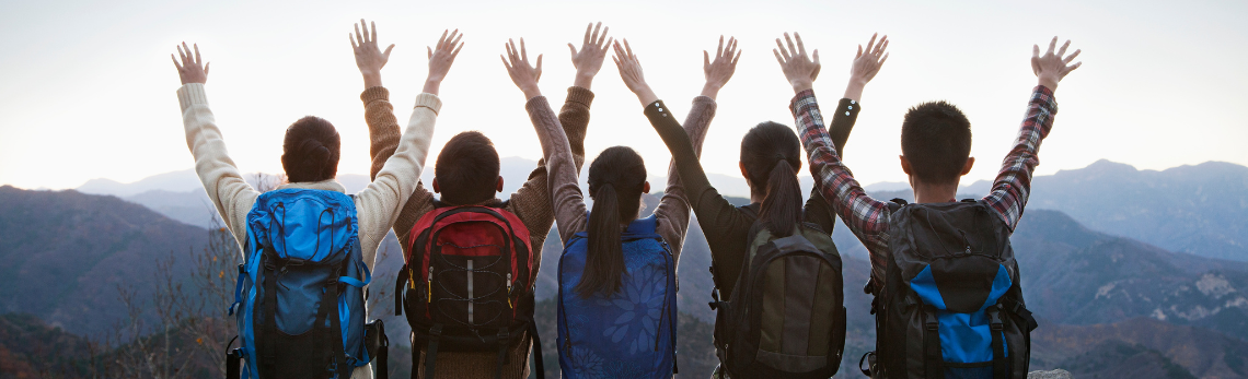 Un groupe de jeunes portant des sacs à dos fait face à une chaîne de montagnes, les bras en l'air.