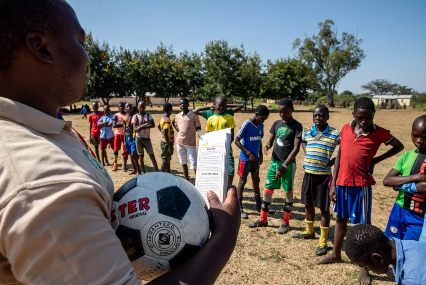 Coach Peter is pictured holding a soccer ball and clipboard. He is standing in front of a group of adolescent boys.